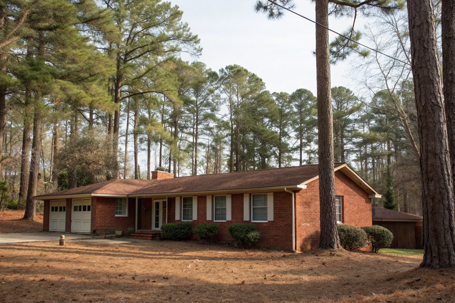 A one-story red brick ranch house with a worn brown shingle roof among tall pines